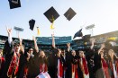 Graduates wearing black and red gowns toss their caps in the air at Fenway Park.