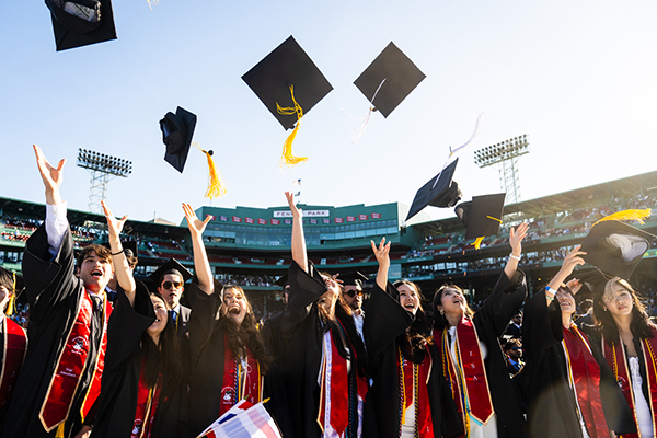 Graduates wearing black and red gowns toss their caps in the air at Fenway Park.