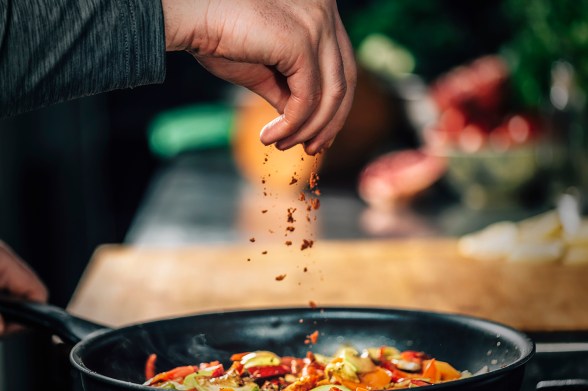 A person's hand sprinkling chili flakes over a pan of food cooking on a stove.