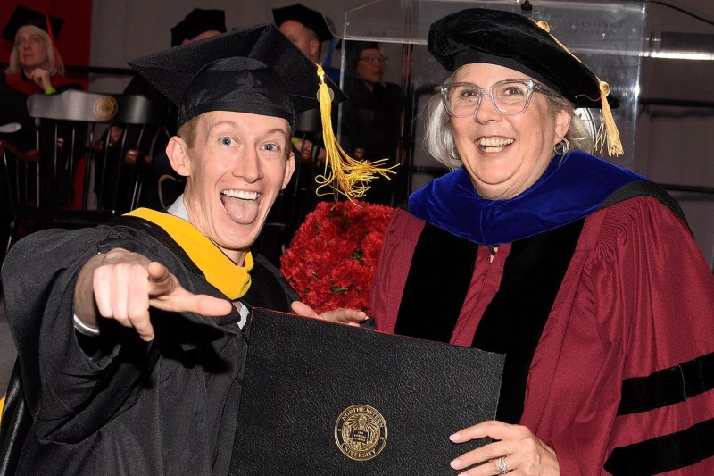 Eric Eldredge posing at his Northeastern graduation ceremony. He has his tongue out and is pointing one hand at the camera. A Northeastern faculty member stands next to him, smiling, while holding his diploma.