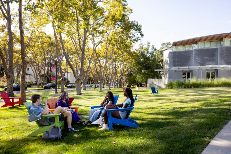 Four students sit on the grass outside the Lokey School on Northeastern’s Oakland campus, talking and relaxing under sunny skies with campus buildings in the background.