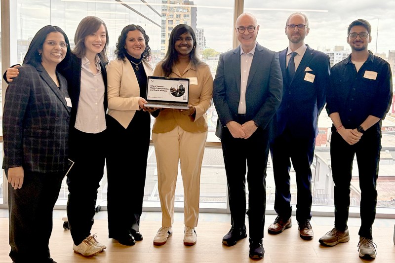A group of students and university administrators dressed in suits hold up a laptop displaying information about Northeastern public transportation research.