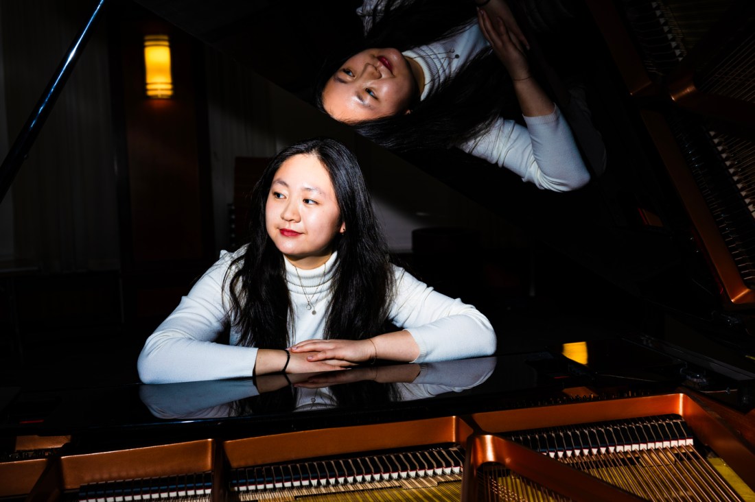 Bonnie Liu sits at a grand piano with the lid open, shot from across the piano so the interior strings and hammers are visible.