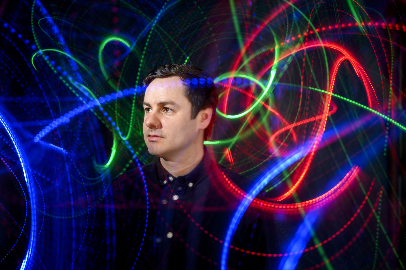 A man in a dark collared shirt looks to the side against a dark background with swirling blue, green and red light trails creating circular and wave patterns around him.