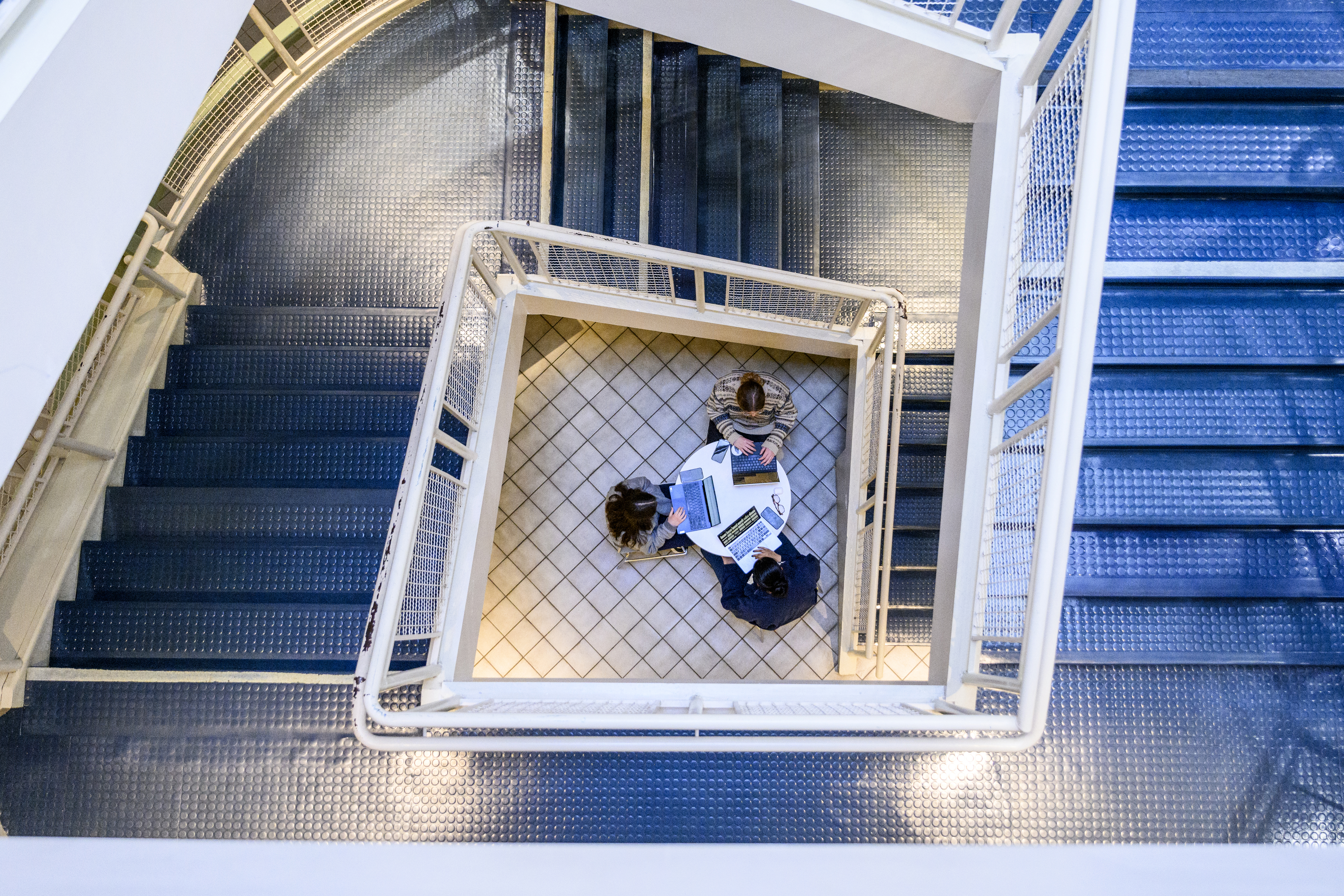 Aerial view of a staircase and three people sitting around a table several floors down. 