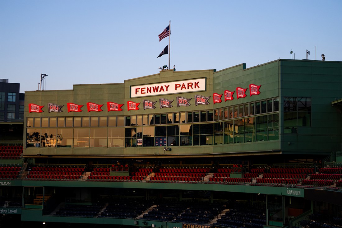 Exterior view of the Pavilion level with the iconic ‘Fenway Park’ sign centered above the seating, championship banners lining the façade, and American and POW/MIA flags flying against a clear sky at sunrise.