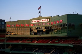 Exterior view of the Pavilion level with the iconic ‘Fenway Park’ sign centered above the seating, championship banners lining the façade, and American and POW/MIA flags flying against a clear sky at sunrise.