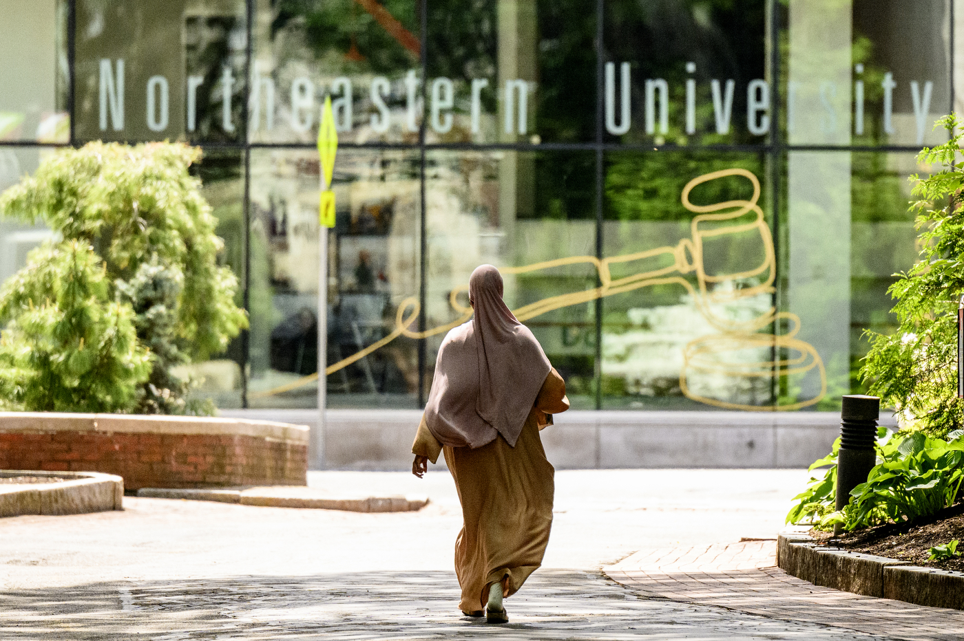 A person wearing a hijab and long garment walks towards a building with glass windows that says 'Northeastern University' on it.