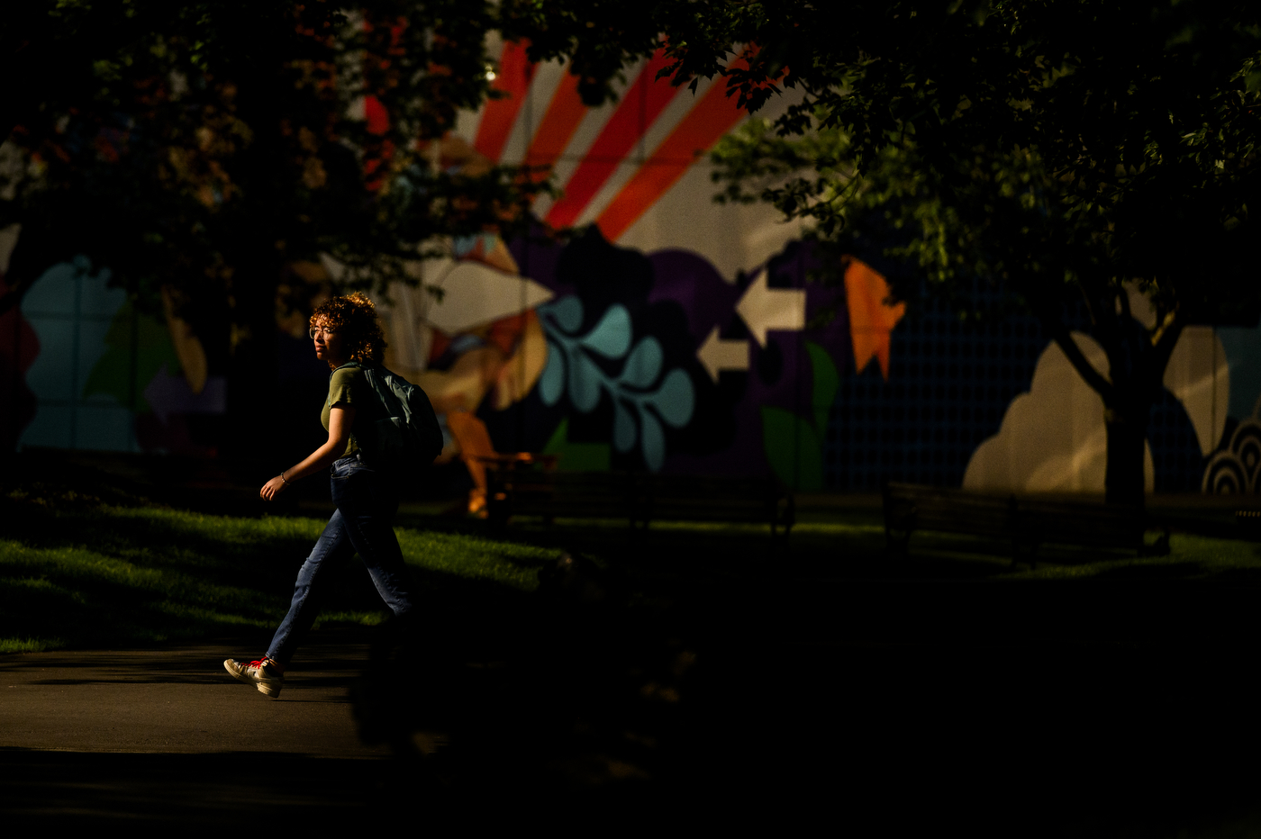 A student walking across campus in front of a wall painted with a mural at nighttime. 