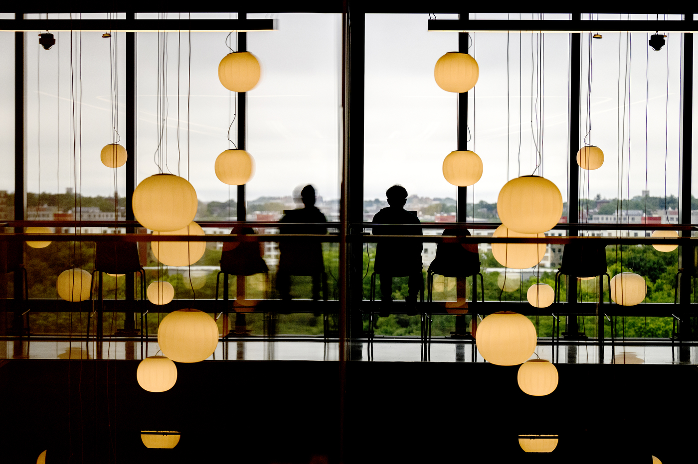 Silhouettes of two people sitting at a counter in front of floor-to-ceiling windows overlooking the Roxbury neighborhood of Boston. Pendant lights hang from the ceiling at various heights.