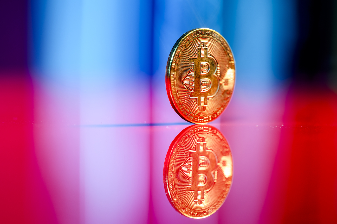 A round gold piece with the Bitcoin symbol on it against a gradient background transitioning from blue to pink to red.