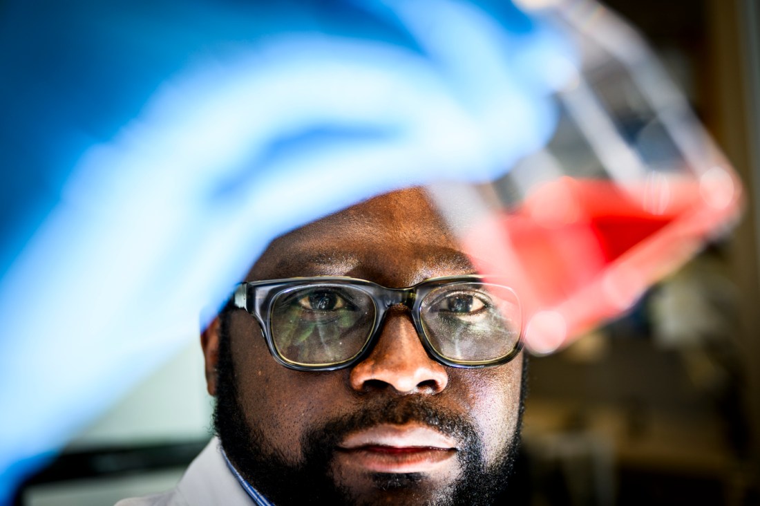 A researcher examines a sample of red liquid in a beaker that they're holding in the foreground.