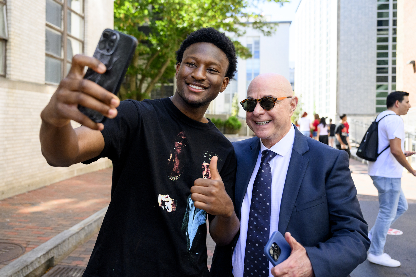 A student in a black shirt takes a selfie with President Aoun wearing a suit and sunglasses on a brick-paved outdoor walkway.