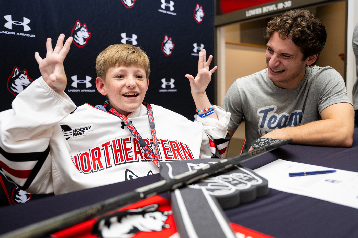 A young boy wearing a Northeastern hockey jersey waves his hands excitedly while sitting at a table with an adult at a signing event. There is a black backdrop with both Under Armor and Northeastern branding on it behind them. 