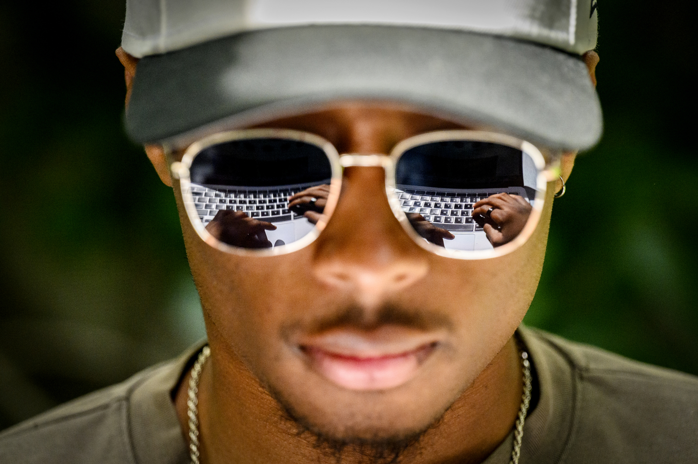 Close-up portrait. ofa person wearing reflective sunglasses and a baseball cap working on a laptop, which is visible in the reflection of his sunglasses.