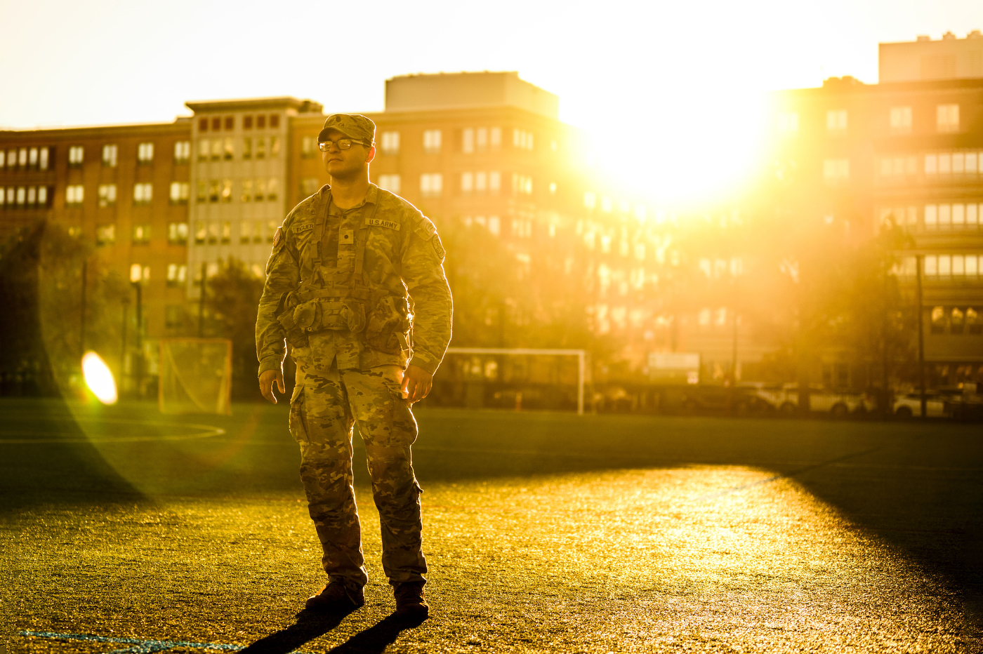 A person in military camouflage uniform stands on Carter Field at sunrise, with residence halls visible in the background.