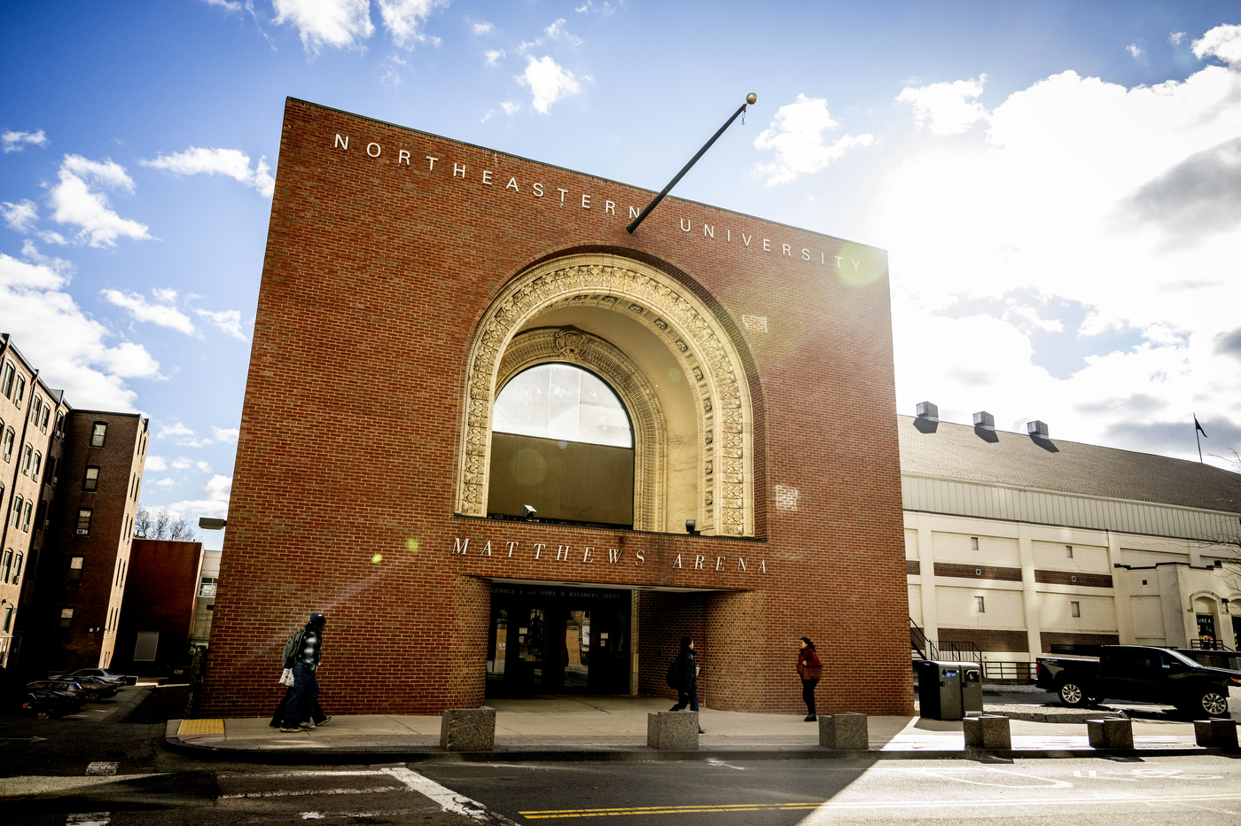 Exterior view of Matthews Arena's decorative brick archway entrance on a sunny day with pedestrians walking outside.