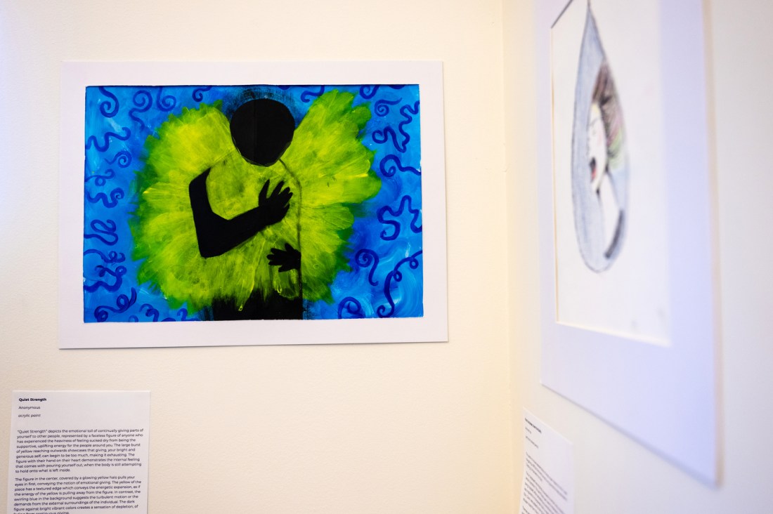 Colorful student artwork hangs on a white gallery wall, including a bright green and blue painting with an abstract human figure.