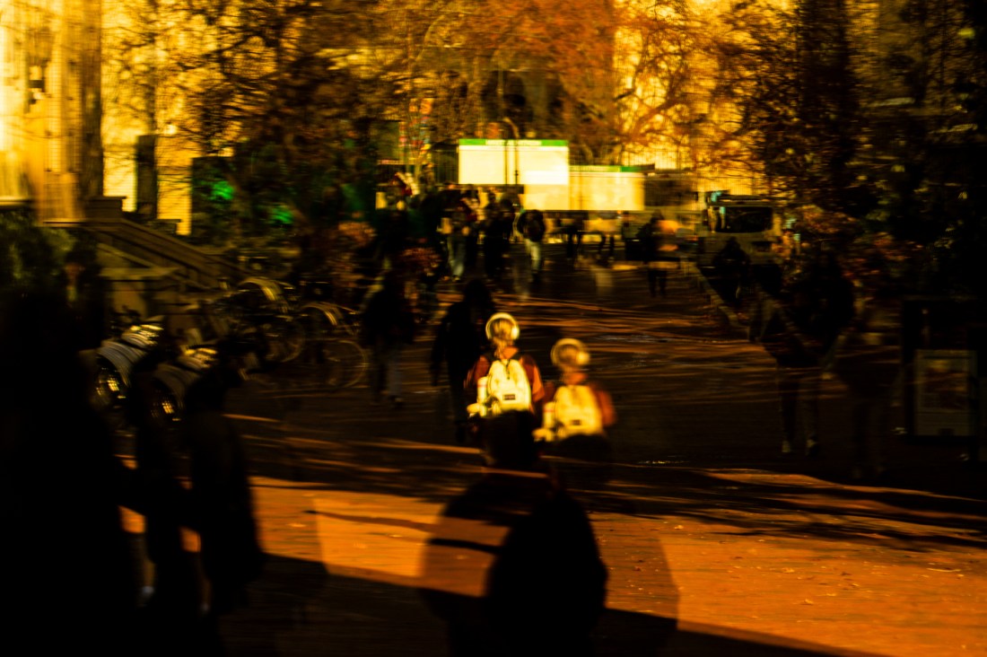 Members of the Northeastern community, as well as signs for the green line, are reflected in glass. The photo has a golden hue. 