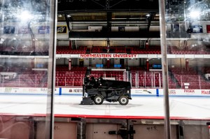 A man drives a black Zamboni ice resurfacing machine inside a hockey rink surrounded by red seats and accents.