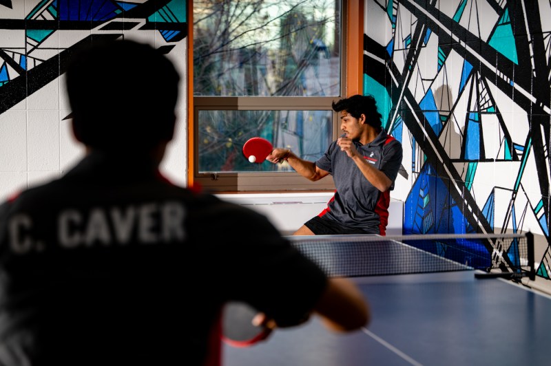 A person hits a table tennis ball while another person awaits the ball on the opposite side of the table. 