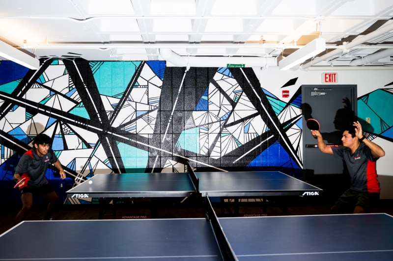 Two people play table tennis in front of an intricate mural. 
