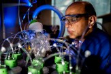 A man with laboratory goggles and a blue suit performs an experiment involving an elaborate system of tubes.