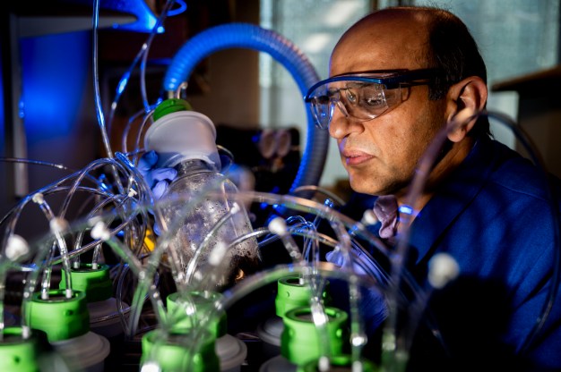 A man with laboratory goggles and a blue suit performs an experiment involving an elaborate system of tubes.