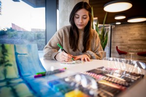 A student with brown hair works on a painting with a green marker.