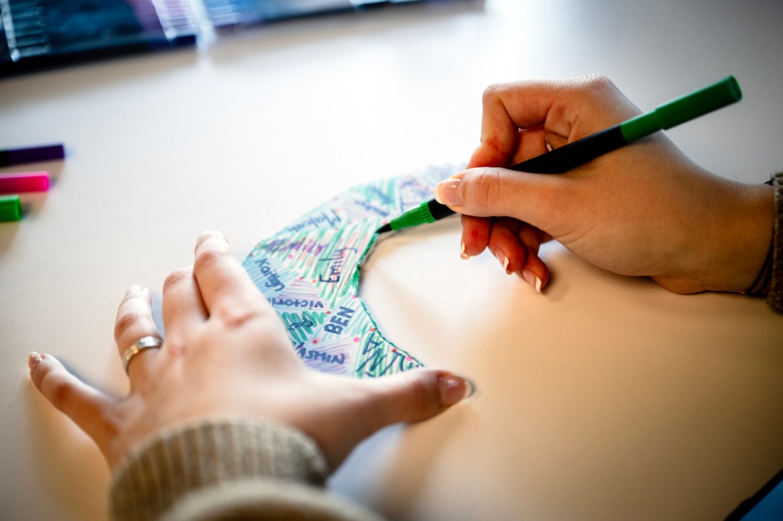 A close-up of a student’s hands coloring a curved paper shape with a green marker at a desk, surrounded by other colored markers.