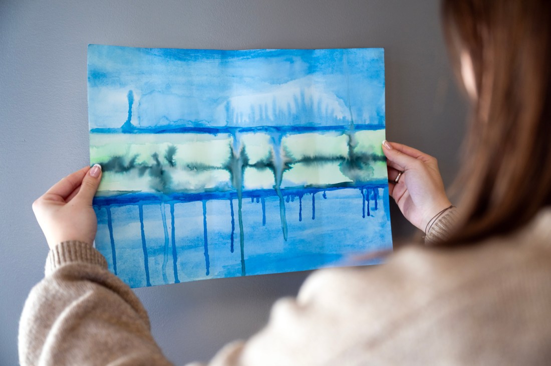 A student holds up a blue watercolor painting with horizontal bands and dripping pigment, viewed from behind against a gray wall.