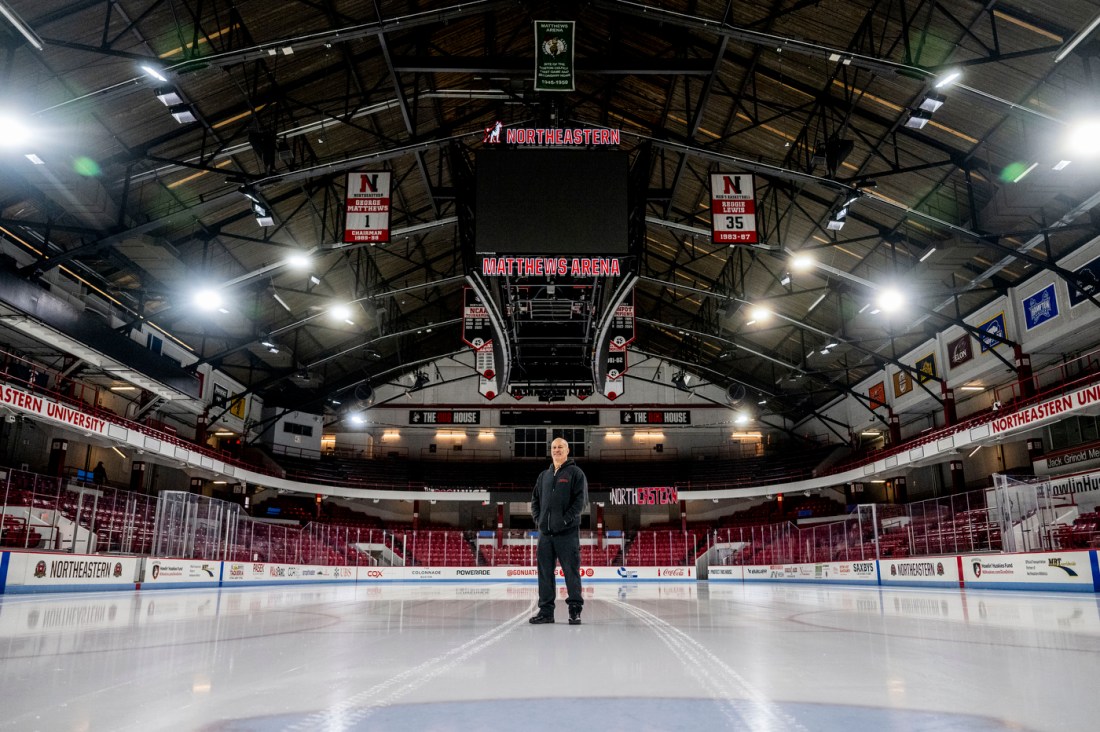 A man smiling and wearing a black zip-up hoodie, black pants and black sneakers stands in the middle of the ice rink under Northeastern banners.