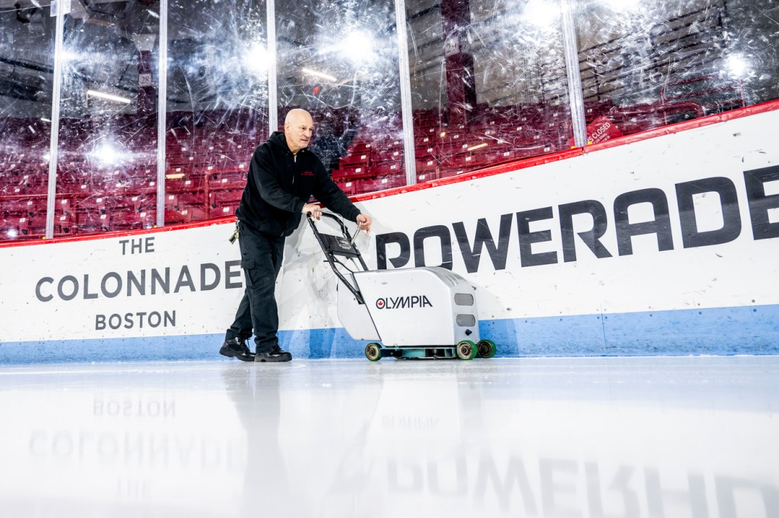 A man wearing a black zip-up hoodie, black walks along the interior edge of the ice rink with a hand-pushed edging machine.