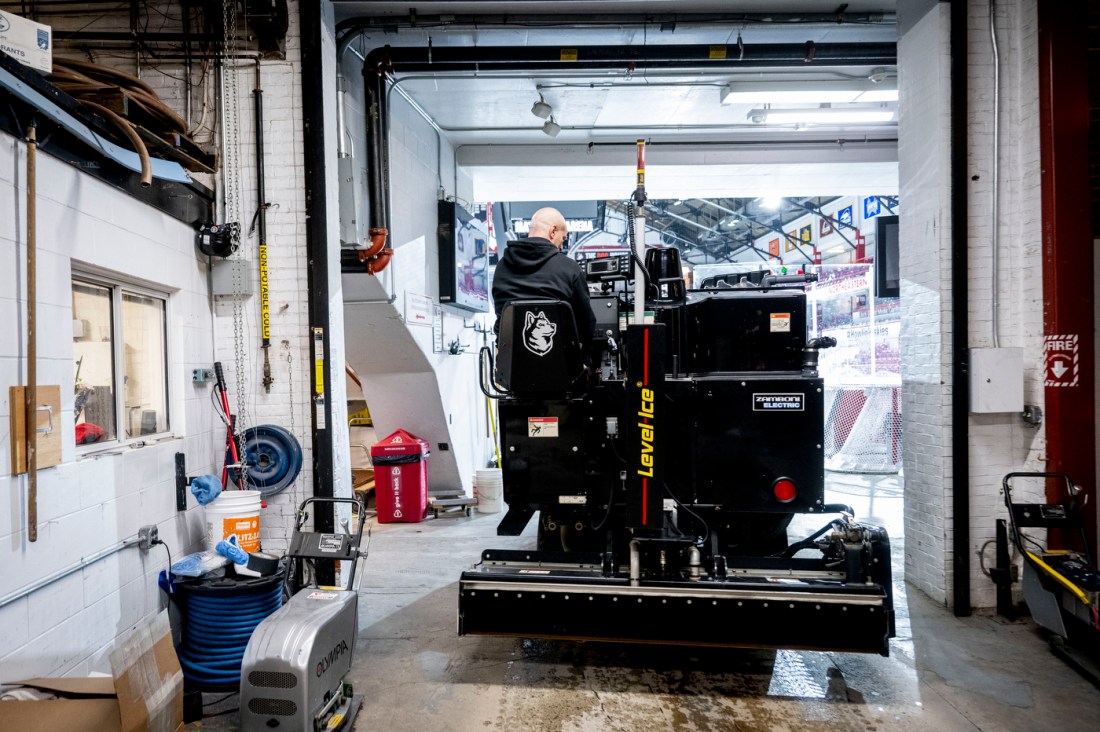 An athletic facilities worker drives an ice-resurfacing machine inside a garage bay at an arena, preparing to enter the rink.