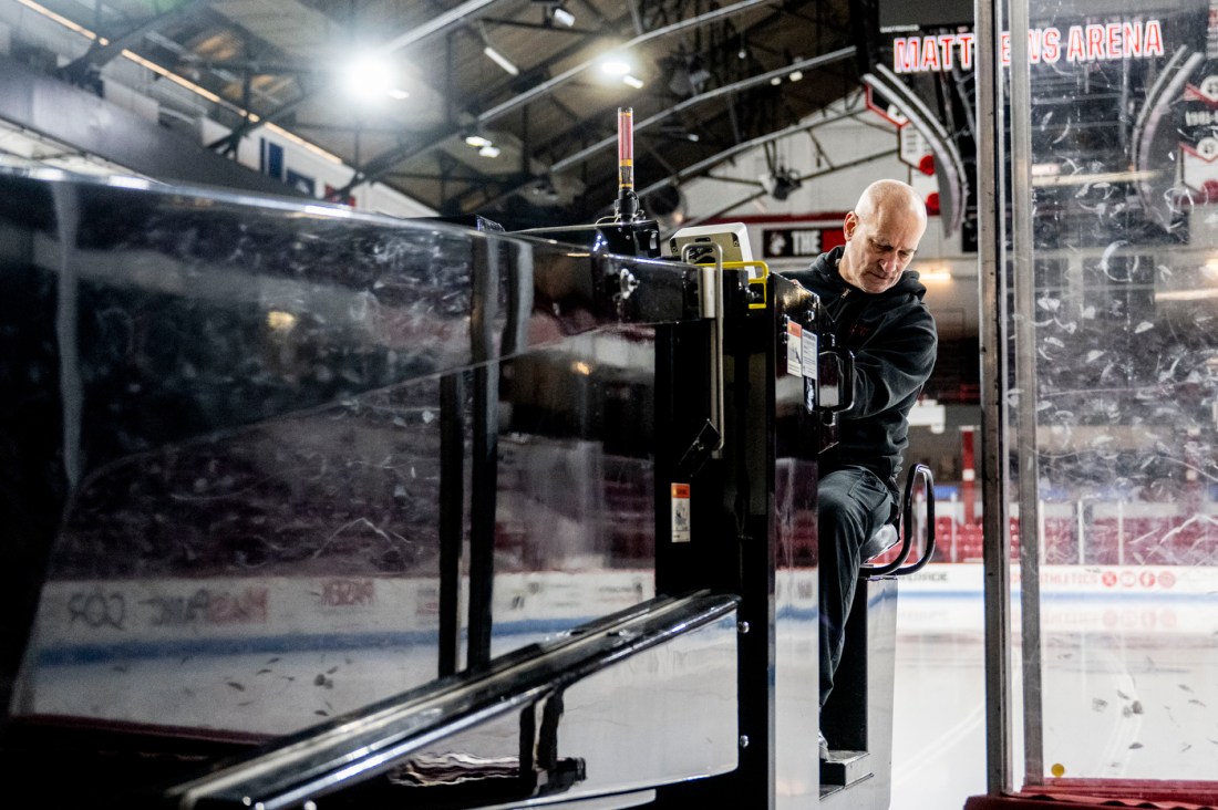 A bald man in black Northeastern hoodie exits ice rink on a black Zamboni machine.