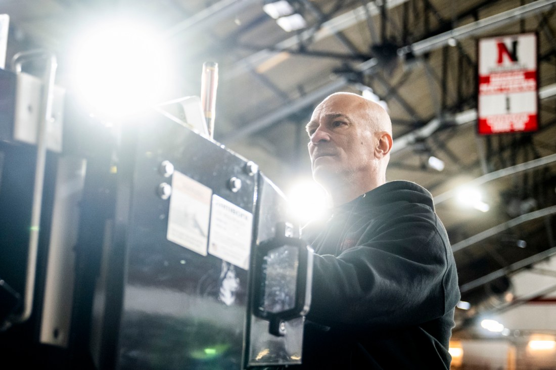 A man in a hoodie works on machinery beneath an ice-resurfacing vehicle inside a maintenance area.