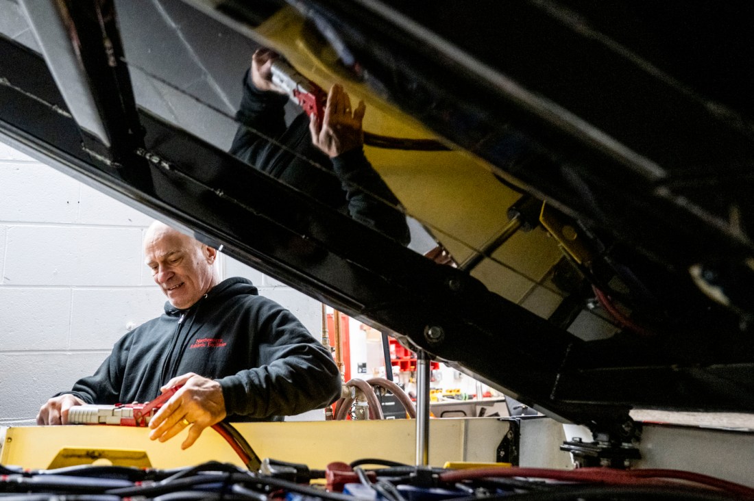 A man in a hoodie works on machinery beneath an ice-resurfacing vehicle inside a maintenance area.