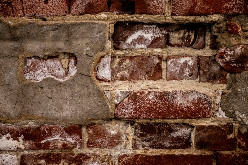 Close-up of weathered red bricks and aged mortar showing white salt deposits and worn texture, with a gray concrete block visible among the brickwork.
