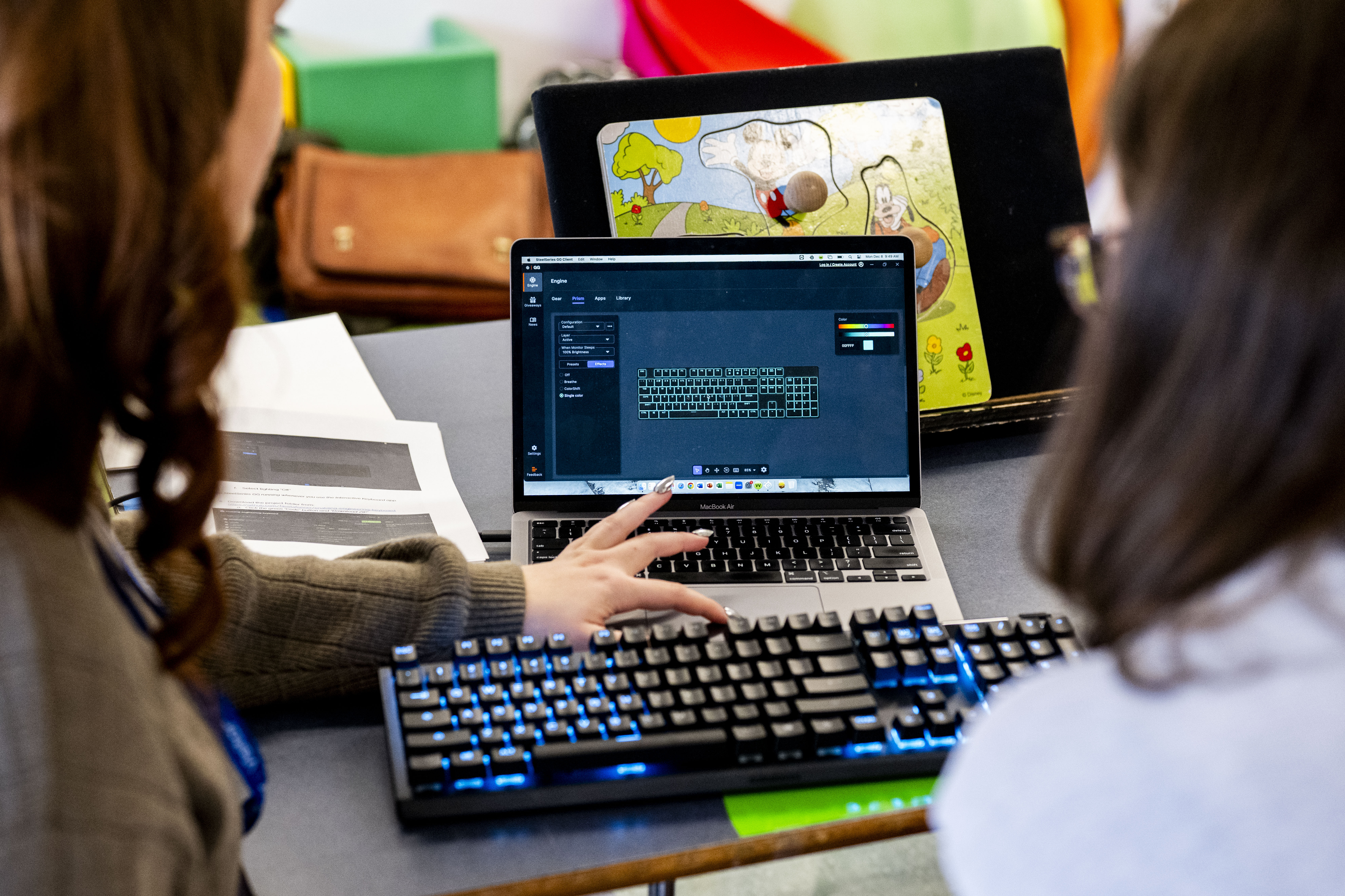 Two students view a tablet displaying an illustrated children's story, with colorful learning materials visible in the background.