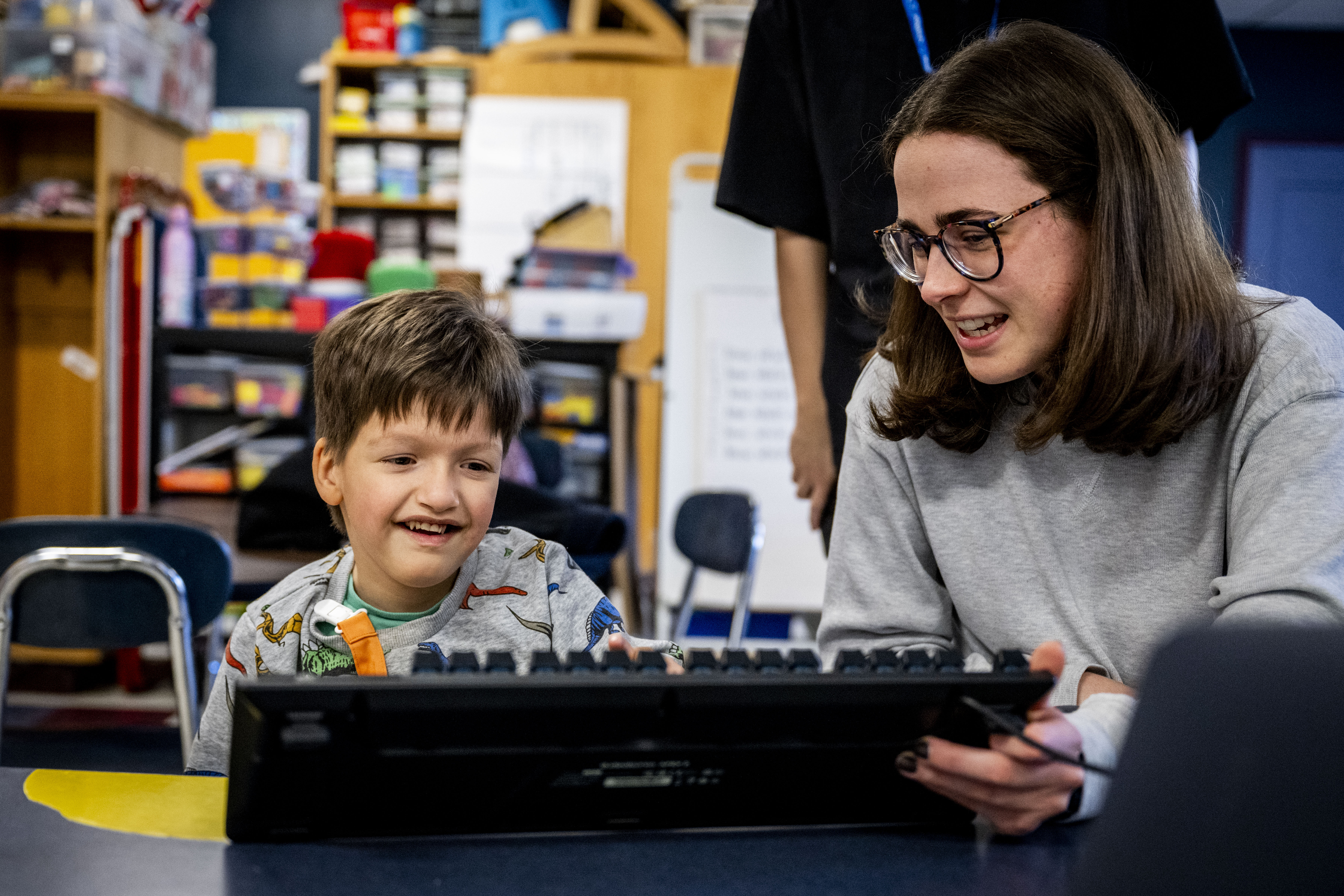 A person with dark hair and glasses in a gray sweater leans over to work with a smiling young student in a dinosaur-print shirt who is seated at a keyboard in a classroom filled with colorful storage bins.
