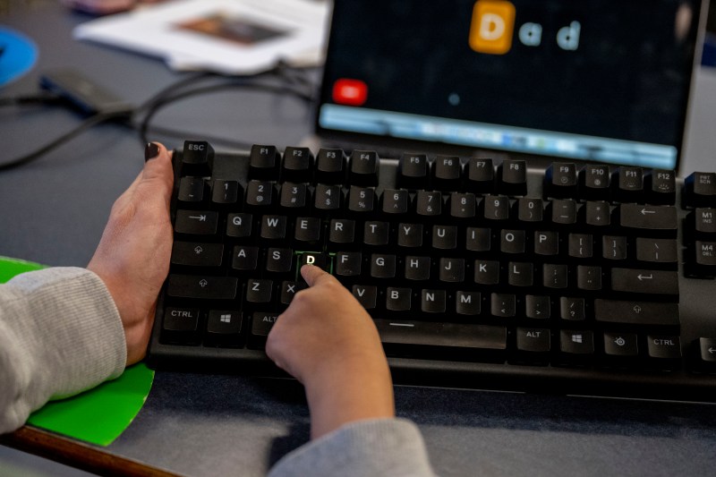 Close up of keyboard with second grade student pressing a key, while a teacher holds it upright.