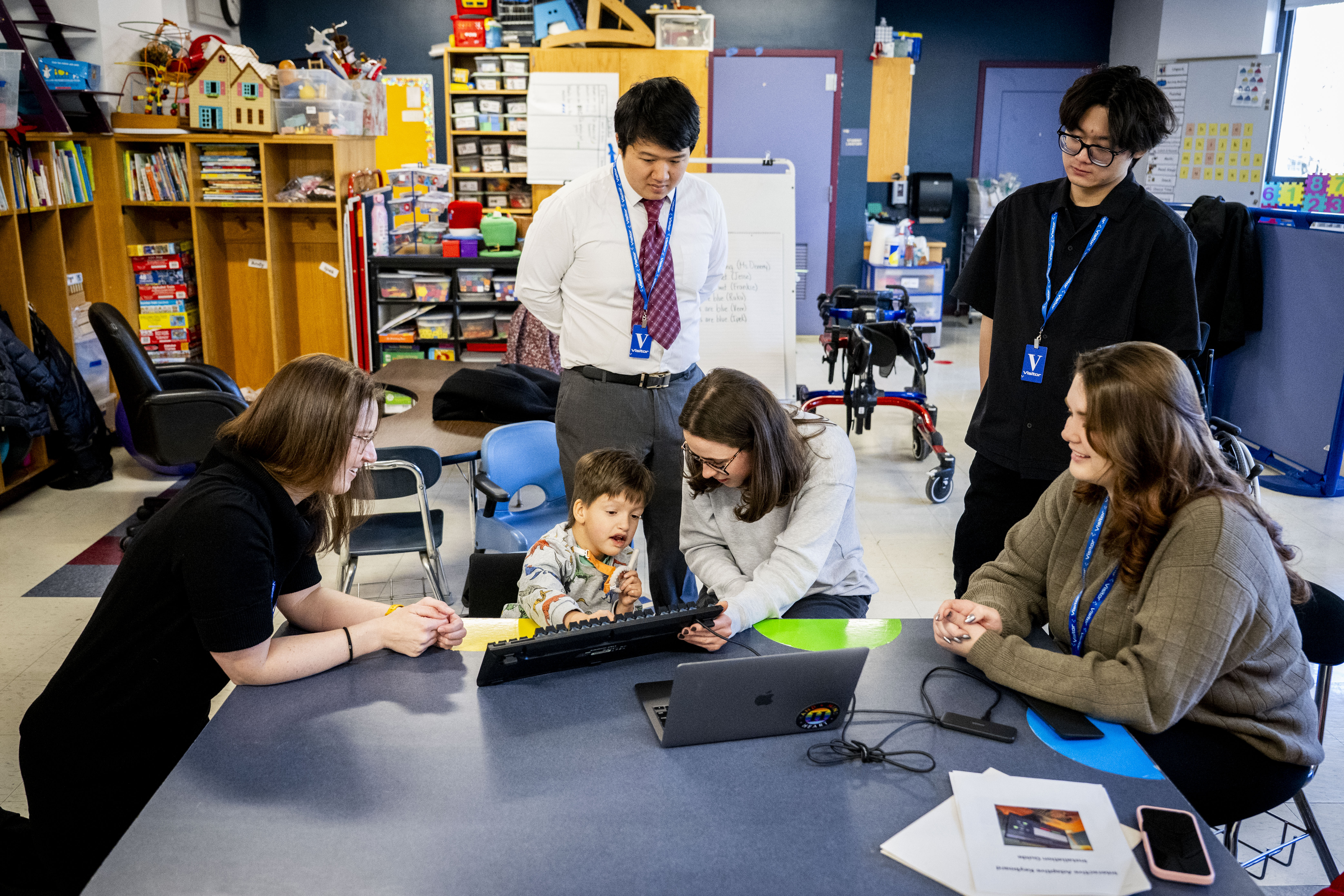 A young student sits at a table surrounded by other people, using an interactive keyboard.