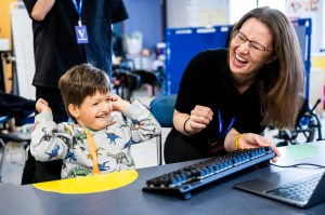 A person with dark hair and glasses laughs joyfully while leaning over a table next to a young student in a dinosaur-print shirt who is seated and smiling. They are working with a black adaptive keyboard with blue accents on the table.