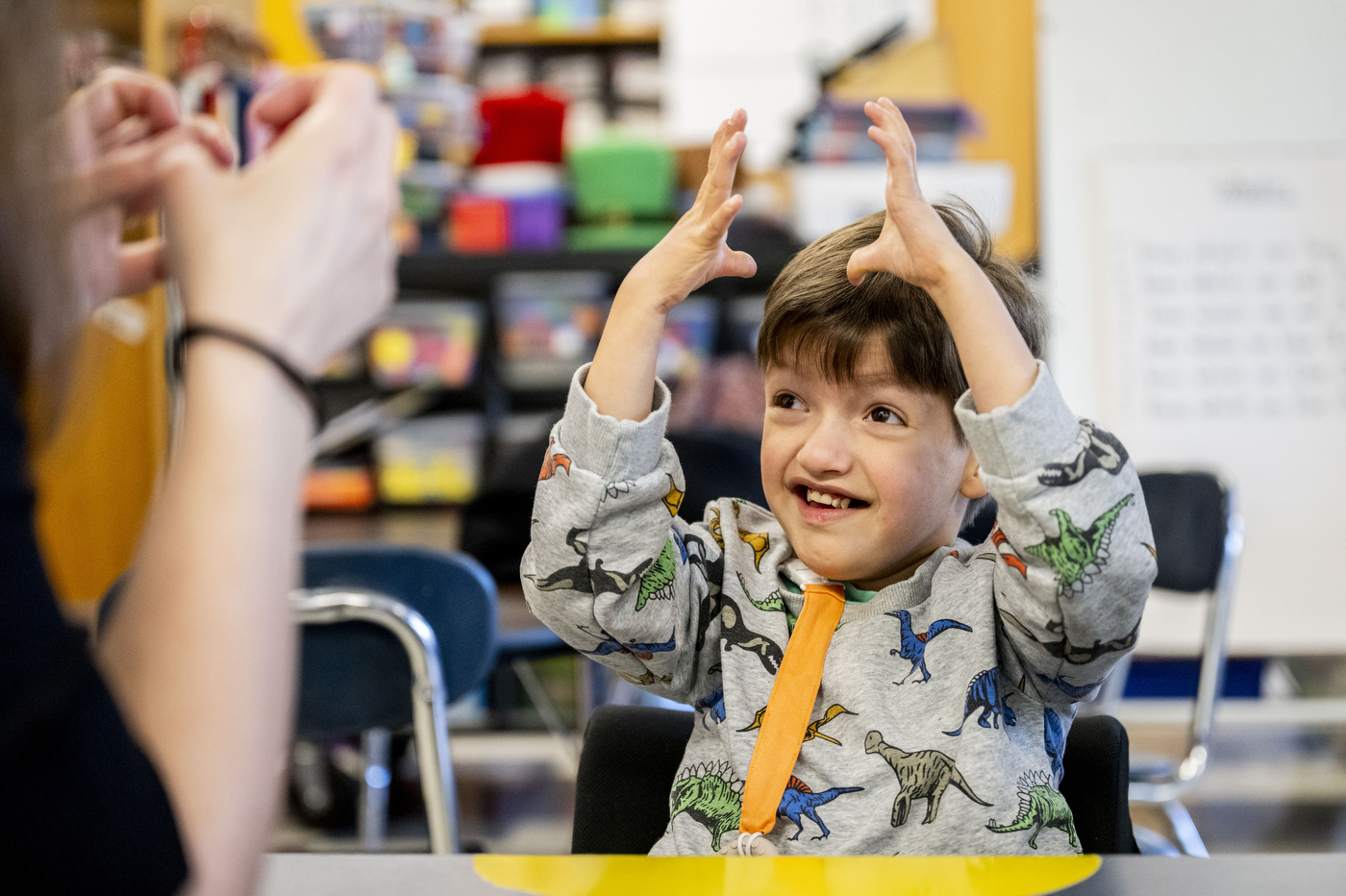 A young student in a dinosaur-print shirt raises both hands above their head in a celebratory gesture while smiling, with colorful classroom materials and learning blocks visible in the background.
