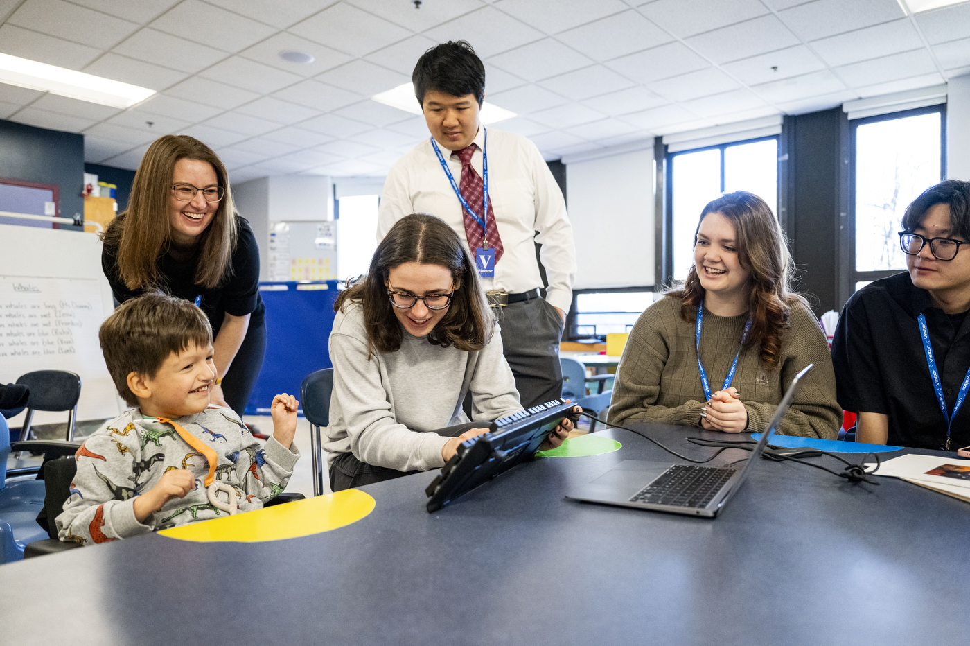 A team of five people gathers around a young student in a dinosaur-print shirt seated at a table with an adaptive keyboard device. The group includes a woman in a black top leaning in from the left, a man in a white shirt and red patterned tie standing in the center, a woman in a gray sweater working with the keyboard, and two additional team members on the right side of the table with a laptop. The bright classroom features white drop ceiling tiles, large windows, and a whiteboard visible in the background.