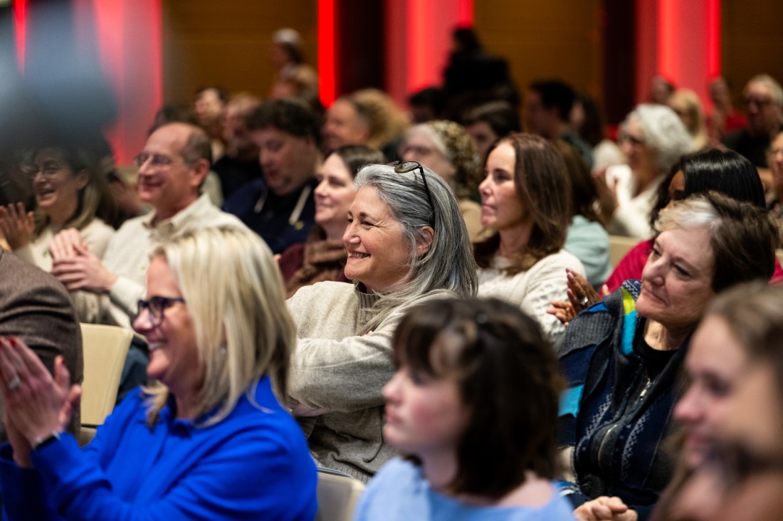 Audience members laugh and applaud during the Morton Ruderman Memorial Lecture at Northeastern University. The diverse crowd is engaged and enjoying the conversation, with red accent lighting visible on the walls in the background creating a warm atmosphere in the venue.