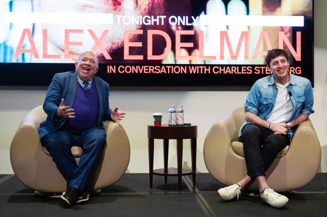 Alex Edelman and Dr. Steinberg sitting on chairs next to each other on a stage in front of a screen that says 'ALEX EDELMAN' on it in pink letters.