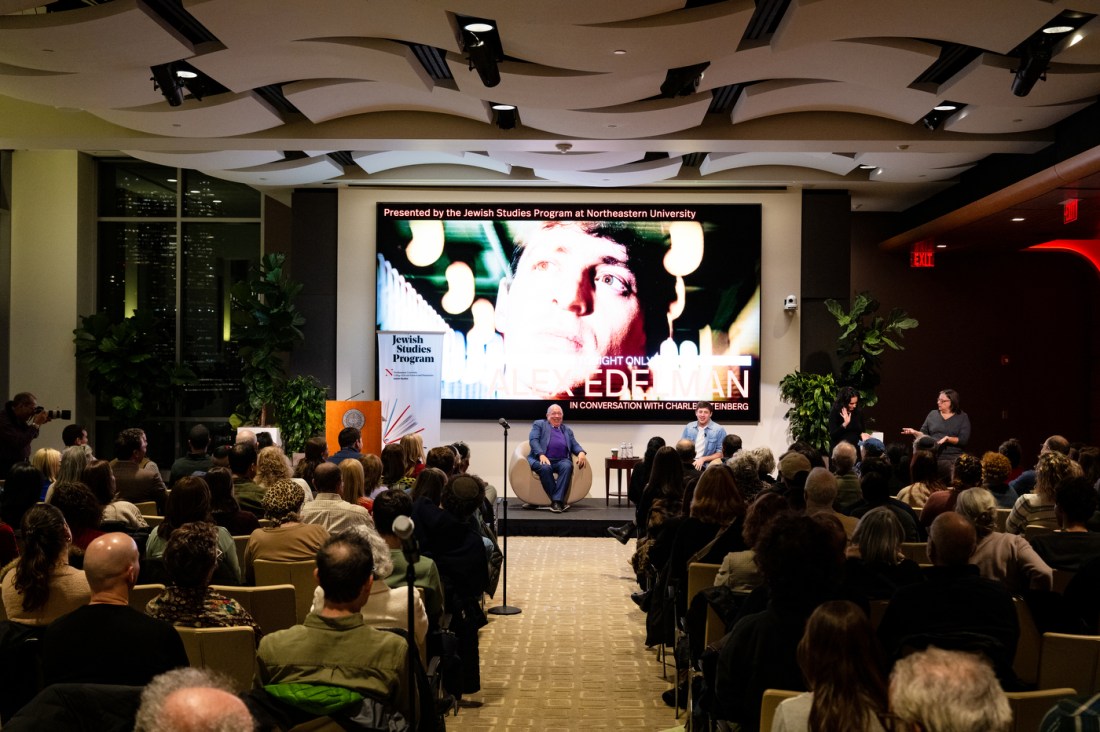 Comic Alex Edelman sits on stage in conversation with Charles Steinberg at Northeastern University's East Village. Behind them, a large screen displays a projection featuring Edelman's face with text reading "Presented by the Jewish Studies Program at Northeastern University" and "Just One: EDELMAN in conversation with Charles Steinberg." The audience sits in rows facing the stage, with plants and modern architectural ceiling details visible in the space.