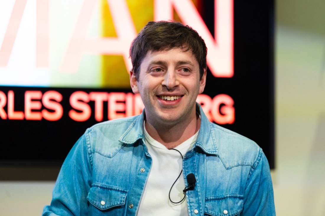 Close-up portrait of comic Alex Edelman smiling warmly at the camera. He wears a light blue denim shirt and is photographed against a backdrop showing partial text reading "RLES STEINBERG" (part of Charles Steinberg's name)