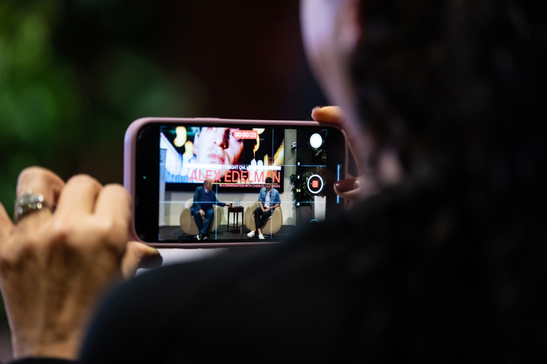 An audience member holds up a smartphone to record the on-stage conversation between Alex Edelman and Charles Steinberg. The phone screen displays the two speakers sitting on stage with "EDELMAN" visible on the projection behind them.